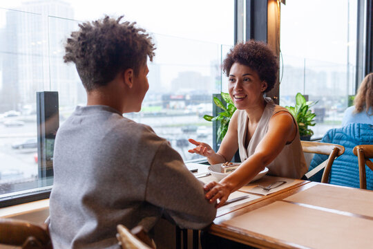 African American woman and her son are sitting in cafe and talking, curly-haired teenager and his mother are sitting in restaurant and smiling