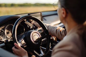 A woman holds the steering wheel while driving on a road. Green fields are visible outside the car. Sunlight comes through the windows, brightening the scene