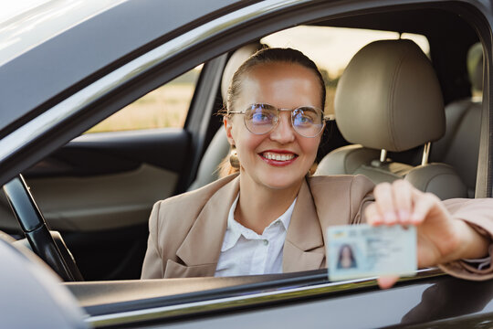 A woman is sitting in a car, smiling and holding up her ID card. The sun is shining and trees can be seen in the background