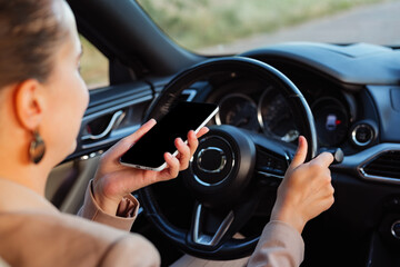 Naklejka premium A woman sits in the driver's seat of a car, holding a smartphone in one hand while steering with the other. The scene is set on a road under clear skies