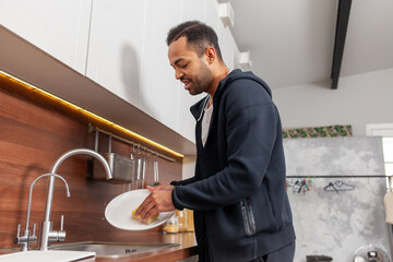African American man washes dishes alone in the kitchen, guy cleans the house and does housework