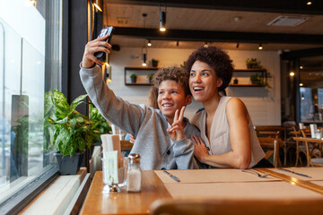 African-American woman and her son are sitting in cafe and taking selfies on smartphone, teenager and his mother are taking photos on phone in restaurant