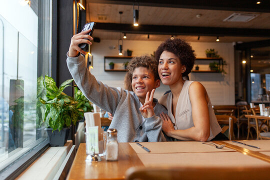 African-American woman and her son are sitting in cafe and taking selfies on smartphone, teenager and his mother are taking photos on phone in restaurant