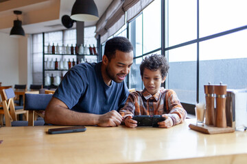 African American man and his son use smartphone in cafe at table, teenager and his father sit in restaurant and play video games on mobile phone