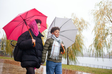 African American woman and her son in down jackets walk in the rain in park with umbrellas, teenage boy walks with his mother outdoors in autumn