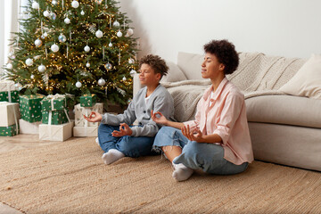African-American woman and her son meditate at home and sit in the lotus position on Christmas Day, while mother teaches teenager to do yoga on New Year's Eve
