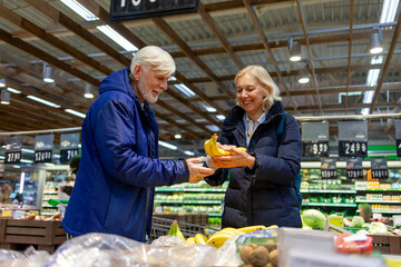 couple of elderly seniors choose products in supermarket, grandparents buy fruits and keep bananas in the store
