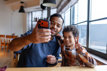 African American man and his son take selfie on smartphone in cafe at table, teenager and his father sit in restaurant and take photo together on mobile phone