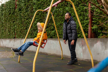 African American man with his son swinging on swing at playground in the city, teenager playing with his father outdoors in autumn