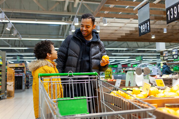 African American man and his son shop for groceries at supermarket together, teenager and his father choose fruit at store and hold orange