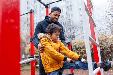 African American man and his son exercise and do exercises outdoors on playground, teenager stretches with his father on exercise equipment