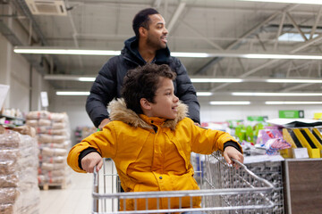 African American man and his son are riding shopping cart in supermarket together, teenager and his father are shopping in store and having fun