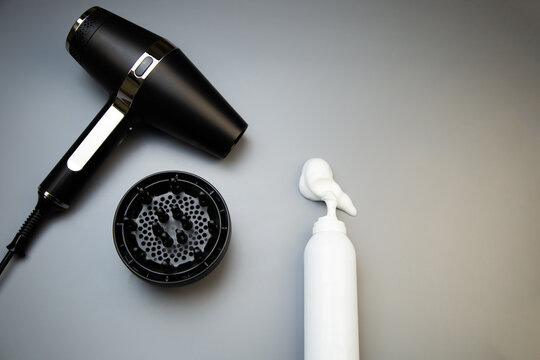 A top view of hair styling foam in a white bottle, next to it is a hairdryer with a diffuser nozzle on a gray background with space for text. The concept of long-term hair styling, creating hairstyles