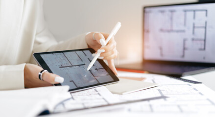 Female interior designer architect analyzing floor plan on digital tablet with laptop and blueprints of apartment