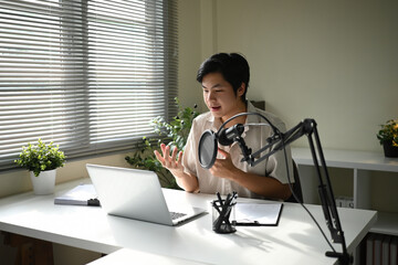 An Asian man records a podcast at home, speaking confidently into a microphone while working on a laptop in a bright, modern workspace