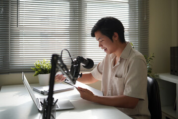 Casual home studio setup with an Asian man hosting an online talk, using a microphone and laptop near a window with natural light