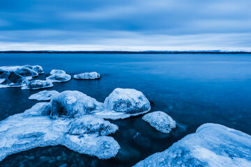 Frozen shoreline at Lake Femunden in Norway shows ice on rocks and still water under cloudy sky