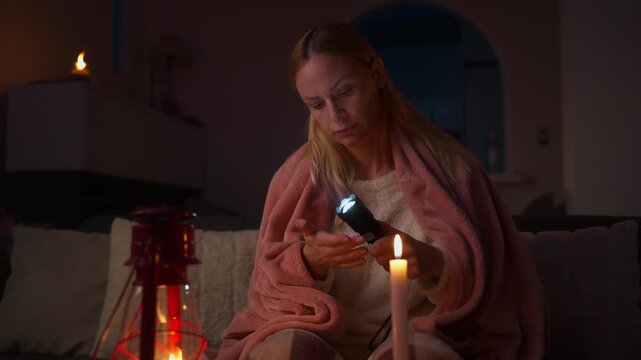 Woman wrapped in blanket sits on sofa during blackout, holding flashlight near face by candle and kerosene lamp, conveying cold, worry and an energy crisis at home
