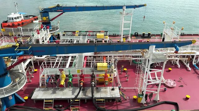 A bunker tanker is refueling other ships at a busy port. Workers are moving equipment and managing hoses on the deck. Many vessels are seen in the background.