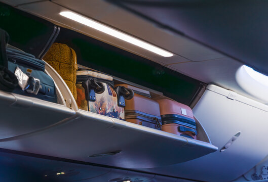 Horizontal wide angle eye level shot of an open overhead bin inside an airplane cabin filled with various travel bags under soft interior lighting