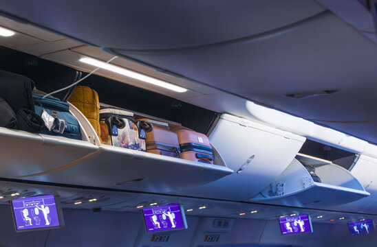Horizontal wide angle eye level shot of an open overhead bin inside an airplane cabin filled with various travel bags under soft interior lighting