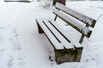 An empty wooden bench with a backrest sits in a winter park, covered with a thick layer of fresh snow. Footprints are visible on the snow-covered ground nearby. Natural daylight, overcast winter day.