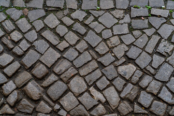 High-angle view of a grey stone pavement made of irregular cobblestones. Wet masonry texture with some green moss in cracks