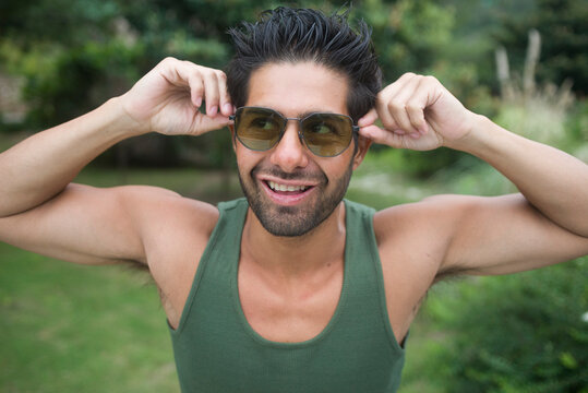 Closeup de hombre joven latino con lentes de sol puestos los sostiene con sus manos de las patillas mientras sonrie