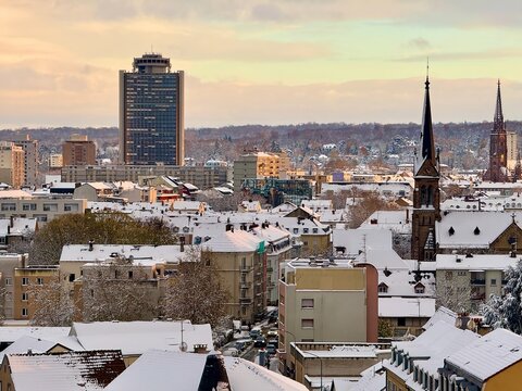 Mulhouse, France - November 21 2024 : Snowy cityscape with Europe Tower high-rise and historic church spires rising above snow-covered rooftops in soft winter sunrise light