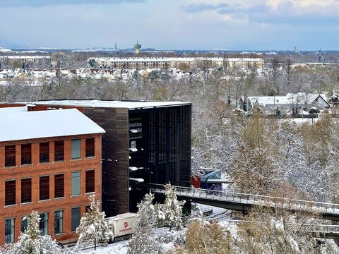 Mulhouse, France - November 21 2024 : Snowy winter view of the National Automobile Museum, showing modern gallery and brick buildings with a footbridge over a river and frosted trees