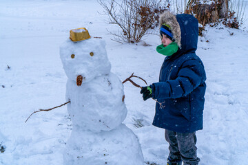 A young child wearing a blue winter jumpsuit, a hat with faux fur, and a green scarf stands next to a handmade snowman. The snowman has a cardboard hat and branch arms. Background shows a blurred wint