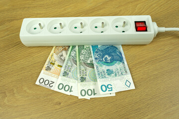 rising electricity prices and high energy costs concept with zloty (PLN) banknotes under white power strip on wooden (oak) background representing household expenses and inflation in Poland 