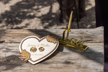 Wedding rings on engraved heart plaque beside golden love pen on rustic wood table