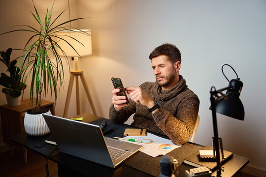 Man sitting at desk using smartphone while working in a home office with laptop and documents. Tired freelancer procrastinating at workplace during remote work. Unproductive man using phone