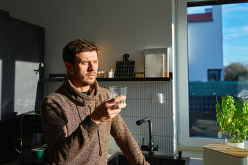 Fototapeta premium Close-up of adult man drinking water from clear glass indoors with natural daylight. Concept of daily hydration, home routine and healthy lifestyle.