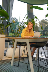 Remote work in coworking space, young woman using laptop and phone, biophilic office interior,...