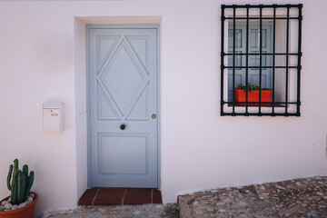 White building wall with light blue door and cactus in terracotta pot nearby. Black window with grid holds red flower pot. Details of Spanish town