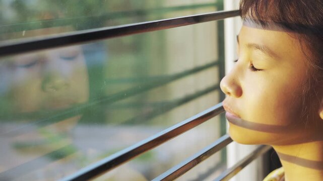 Sad Asian girl looking out window through iron bars during sunset. Lonely child waiting with hope, sun shadows on face and reflection on glass. Concept of depression, solitude, or home isolation.