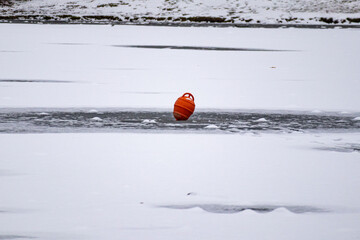 Red plastic buoy or container standing on frozen water surface surrounded by snow.