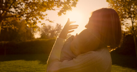 A cheerful woman carries her baby while walking through a park during sunset, sharing loving...
