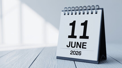 Minimalist standing desk calendar, bold black 11 June 2026 on white page, spiral binding, light gray wooden texture, soft blurred blue-gray background, angled shadow, modern clean aesthetic.