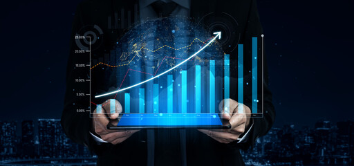 A businessman stands in a dark urban environment, holding a tablet displaying digital graphs and financial data, illustrating growth and success in modern finance. Scalp