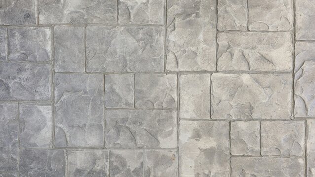 A detailed close-up of a grey stamped concrete surface, designed with an irregular stone block pattern. The texture is rough and natural, providing a seamless background of a paved floor or walkway.