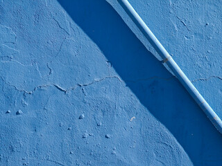 Textured blue wall with a white pipe casting a shadow. The surface shows signs of wear and unevenness, creating an interesting visual effect.