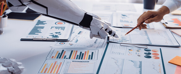 This image captures a robotic arm working alongside a human hand over various business charts and graphs in a bright office environment, showcasing modern automation.Noogenesis