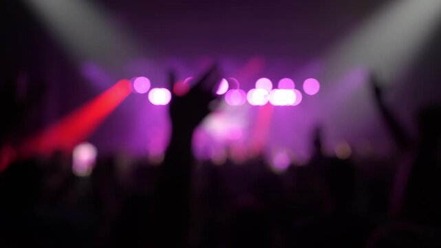 Abstract blurred concert scene with purple stage lights and red spotlight beams. A large audience appears as dark silhouettes in front of the stage.