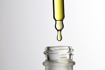 Studio shot of a glass pipette with a hanging drop of golden cosmetic oil above an open glass bottle, isolated on a white background
