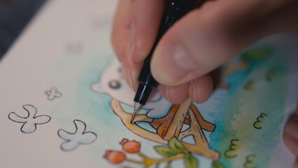 Artist fingers with ink pen drawing details on miniature watercolor painting. Ghost cat on garden bench. Macro shot, selective focus on pen and paper, blurred background, whimsical fairytale process.