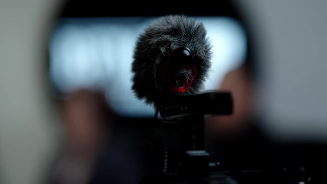 Close-up of a shotgun microphone with furry windscreen mounted on a camera rig during an indoor interview setup, capturing dialogue with shallow depth of field and studio lighting.