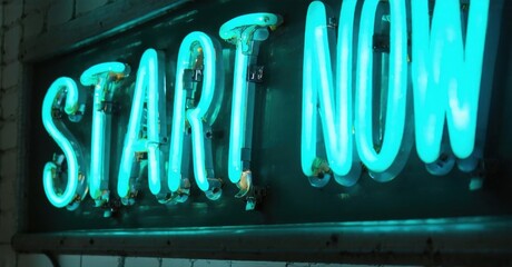 Bright neon sign displays words Start Now. Turquoise glow contrasts dark background. Simple message, motivational impact.